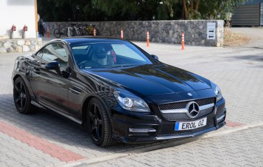 Gaziveren Cyprus -05.12.2024 A stylish black sports car stands on a cobblestone street under a clear sky, surrounded by trees and quiet surroundings. clipart