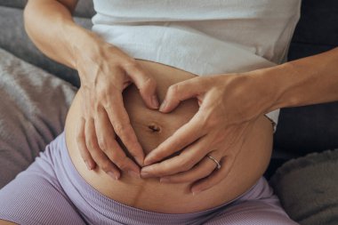 Hamile kadın göbeğinde parmaklarıyla kalp şekli yapar..