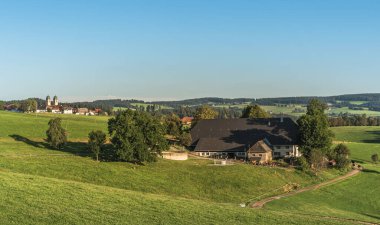 Traditional farmhouse in the Black Forest, surrounded by green meadows and trees, with famous monastery in the background, St. Maergen, Baden-Wuerttemberg, Germany clipart