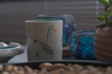 Zarif bir şekilde tasarlanmış, mavi mozaik mum tutucuları, bir seramik kap ve sulu bir bitki tarafından çevrelenmiş çiçek kupası içeren sıcak ve davetkar bir natürmort kompozisyonu.. 