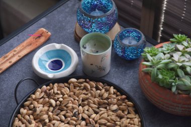 Kavrulmuş şam fıstığı, mavi mozaik mum tutacağı, seramik kül tablası, çiçek balmumu kabı, ahşap tütsü ocağı ve terasotta kabında sulu bir bitki, sıcak ve kırsal bir titreşim yaratıyor..