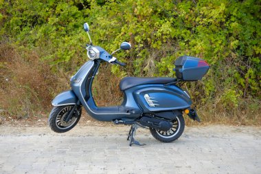 Çevre dostu ulaşımı, şehir yaşam tarzını, hareketliliği, özgürlüğü, rahatlığı ve çağdaş seyahat kavramını temsil eden yeşil yapraklı arka planda park edilmiş modern mavi scooter..