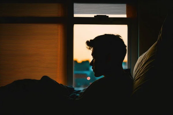 A man sitting in a bed looking out a window at the sunset or sunrise with his hair blowing in the wind backlighting a stock photo neoplasticism