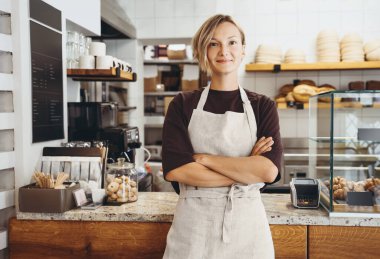 Smiling female baker entrepreneur standing at the counter of bakery and coffee shop. Young woman in cafe near showcase with fresh croissants and bread. Local small business owner indoors.