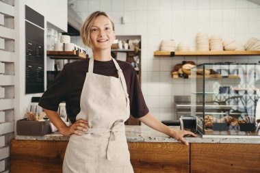 Smiling female baker entrepreneur standing at the counter of bakery and coffee shop. Young woman in cafe near showcase with fresh croissants and bread. Local small business owner indoors.
