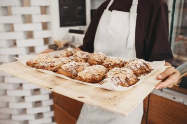 Welcoming female baker holding freshly baked almond croissants in  background of bakery. Young smiling seller woman in cafe or coffee shop. Bistro owner indoors. Sustainable local small business.