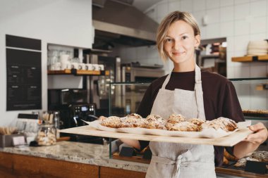 Welcoming female baker holding freshly baked almond croissants in  background of bakery. Young smiling seller woman in cafe or coffee shop. Bistro owner indoors. Sustainable local small business.