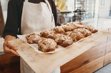 Welcoming female baker holding freshly baked almond croissants in  background of bakery. Young smiling seller woman in cafe or coffee shop. Bistro owner indoors. Sustainable local small business.