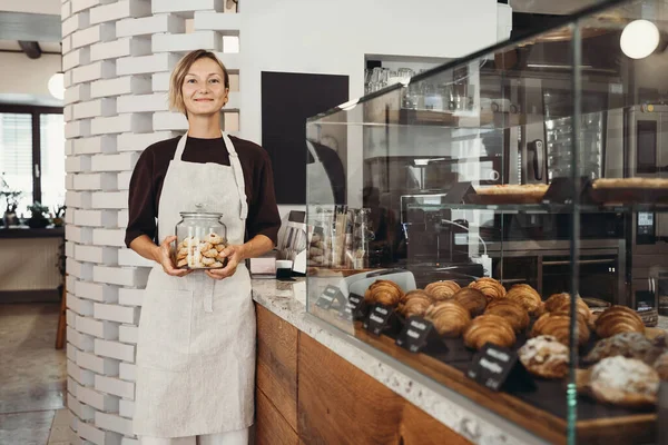 Smiling female baker entrepreneur standing at the counter of bakery and coffee shop. Young woman in cafe near showcase with fresh croissants and bread. Local small business owner indoors.