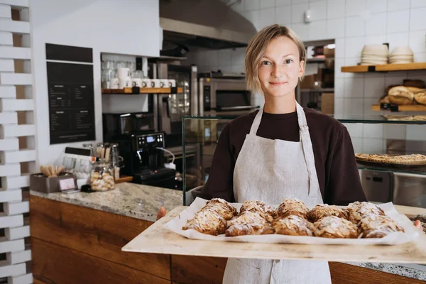 Welcoming female baker holding freshly baked almond croissants in  background of bakery. Young smiling seller woman in cafe or coffee shop. Bistro owner indoors. Sustainable local small business.