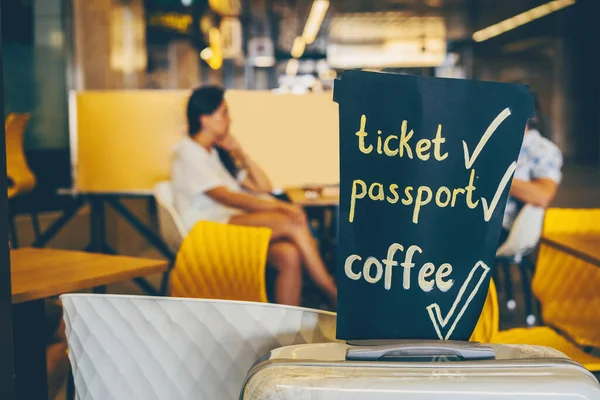 Travel suitcase with black board with list for traveler: tickets, passport, coffee - interior decor in cafe in background with visitors at the airport or railway station. Travel concept.