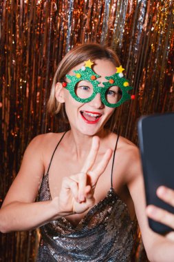 Mutlu kadın akıllı telefondan yeni yılı kutlarken selfie çekiyor. Güzel bir kadın yeni yıl partisinde eğleniyor. Tatil kutlaması. Genç kız mutluluk ve şaşkınlık duygularını ifade eder..
