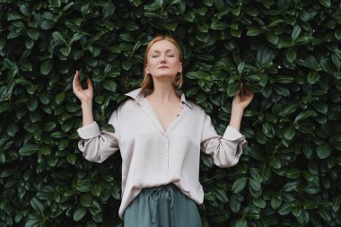 Yeşil çitin arka planında güler yüzlü çekici bir kadın. Yeşil yaprakların önündeki mutlu, kendine güvenen kadın. Neşe Kavramı, Zen ve Denge İnsanları. Ruh sağlığı yoluyla istikrar.