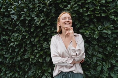 Yeşil çitin arka planında güler yüzlü çekici bir kadın. Yeşil yaprakların önündeki mutlu, kendine güvenen kadın. Neşe Kavramı, Zen ve Denge İnsanları. Ruh sağlığı yoluyla istikrar.