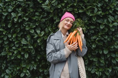 Yeşil yaprak duvarında taze organik havuç ve ağ torbasıyla gülümseyen kadın. Eko yerel ürünleri plastik ambalajlar olmadan sürdürülebilir alışveriş yapma kavramı. Minimalist yaşam tarzı