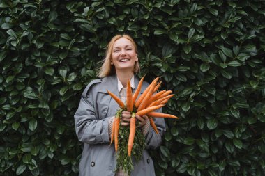Yeşil yaprak duvarında taze organik havuçlarla gülümseyen kadın. Sürdürülebilir alışveriş kavramı, yerel ekolojik ürünler, plastik ambalaj yok. Diyet beslenme, vitaminler. Minimalist vejetaryen yaşam tarzı