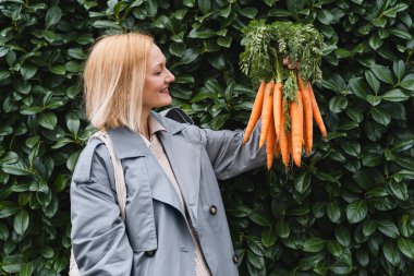 Yeşil yaprak duvarında taze organik havuçlarla gülümseyen kadın. Sürdürülebilir alışveriş kavramı, yerel ekolojik ürünler, plastik ambalaj yok. Diyet beslenme, vitaminler. Minimalist vejetaryen yaşam tarzı