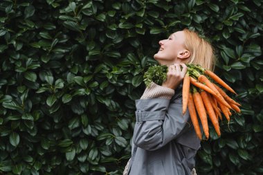 Yeşil yaprak duvarında taze organik havuçlarla gülümseyen kadın. Sürdürülebilir alışveriş kavramı, yerel ekolojik ürünler, plastik ambalaj yok. Diyet beslenme, vitaminler. Minimalist vejetaryen yaşam tarzı