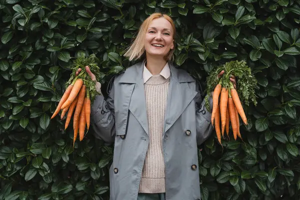 Smiling woman with fresh organic carrots against green leaf wall. Concept of sustainable shopping, eco local products, no plastic packaging. Diet nutrition, vitamins. Minimalist vegan lifestyle