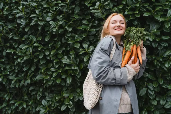 Smiling woman with fresh organic carrots against green leaf wall. Concept of sustainable shopping, eco local products, no plastic packaging. Diet nutrition, vitamins. Minimalist vegan lifestyle