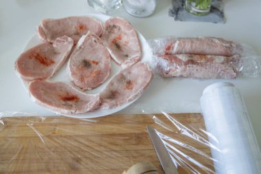 Etleri pirzola için hazırlıyorum. Parçalara ayrılmış çiğ domuz pirzolası ve yapışkan filmlerle kaplı baharatlar serpiştirilmiş..