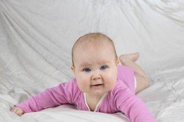 Sevimli ve güzel 4 aylık bir kız bebek yatakta uzanıyor ve kameraya bak. Beyaz arka planda pembe tulumlu bir bebek..