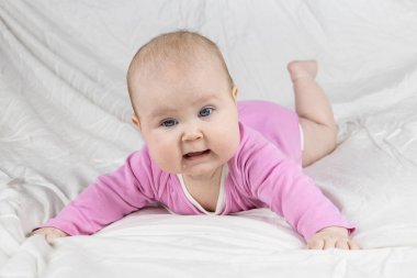Sevimli ve güzel 4 aylık bir kız bebek yatakta uzanıyor ve kameraya bak. Beyaz arka planda pembe tulumlu bir bebek..