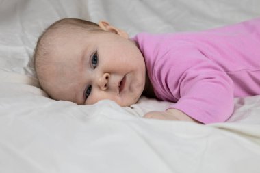 Sevimli ve güzel 4 aylık bir kız bebek yatakta uzanıyor ve kameraya bak. Beyaz arka planda pembe tulumlu bir bebek..