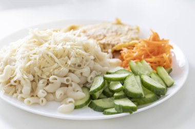 Peynirli makarna, taze salatalık ve havuç ve beyaz tabakta domuz pirzolası.