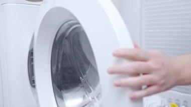 Woman loads colorful clothes into drum stock of washing machine. Household chores in laundry room.