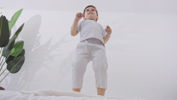 寝室の白い新鮮な寝具が付いているベッドで跳ぶかわいい男の子 家で一人で遊ぶ楽しそうな笑顔の子供 スローモーション — ストック動画