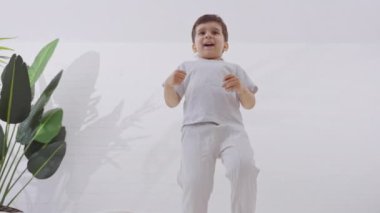 Sevimli küçük çocuk beyaz yatak örtüsüyle yatakta zıplarken eğleniyor. Sağlık, mutluluk ve gençlik kavramı. Yavaş çekim. 
