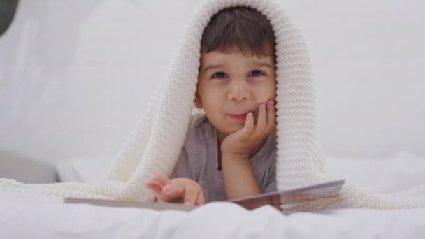 可爱的笑着的小男孩看着一本书 学龄前儿童躺在床上看故事书中的图画 白人幼儿在家里的床上看书 在摄像机前观看 — 图库视频影像