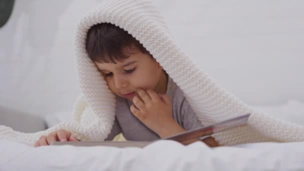 可爱的小男孩在看一本书 学龄前儿童躺在床上看故事书中的图画 托德勒在家里的床上看书 — 图库视频影像