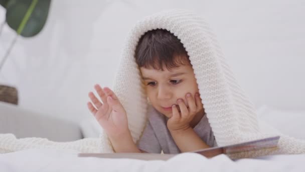 可爱的白人小男孩在看一本书 学龄前儿童躺在床上看故事书中的图画 托德勒在家里的床上看书 — 图库视频影像
