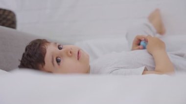 Sevimli küçük bir çocuk beyaz temiz bir yatak ile yatakta dinlenerek uyanıyor. Mutlu çocuk evde oyun oynuyor. Ağır çekim. Uzayı kopyala Çocukluk kavramı.