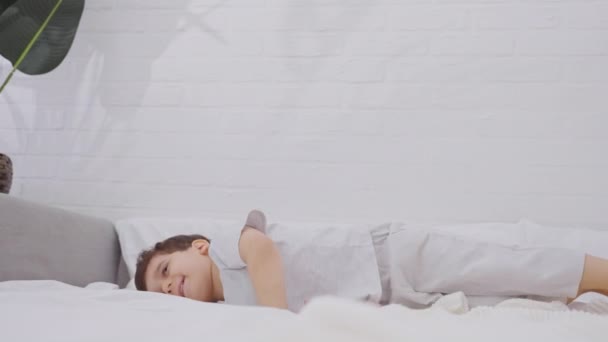 可爱的小男孩在床上嬉闹着 床上铺着洁白新鲜的床上用品 笑得开心的孩子鬼混 一个人在家里玩 慢动作 — 图库视频影像