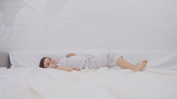 可爱的小男孩醒来后 带着洁白新鲜的床上用品在床上放松 快乐的孩子鬼混 一个人在家里玩 慢动作 — 图库视频影像