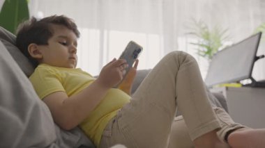 Neşeli çocuk evdeki kanepede oturup video oyunu oynarken cep telefonuna dokunuyor, modern alet edevat akıllı telefon kullanıyor, film izliyor, eğleniyor, oturma odasında dinleniyor.