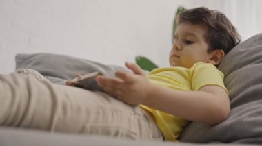 Anaokulu çocuğu seyyar ekrana bakıyor, modern cihaz akıllı telefon kullanmayı, sosyal medya uygulamalarını izlemeyi ya da online oyun oynamayı seviyor, evdeki koltukta oturuyor.. 