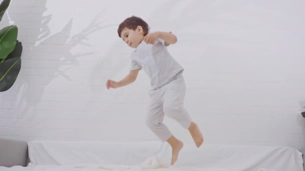 兴奋的小男孩高兴地跳着 倒在白色的新鲜床上用品上 快乐的孩子在家里跳着跳着玩着有趣的活动 自由和幸福童年的概念 慢动作 — 图库视频影像
