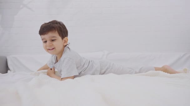 カメラを見ているかわいい笑顔の少年は 両親の寝室の白い寝具にあります 幸せな子供の子供の閉鎖 幸福と自由と幸せな子供時代の概念 — ストック動画