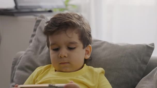 フロントビューの小さな子供の男の子は モバイル画面を見て 漫画を見て 楽しみを持って モバイルオンラインゲームをプレイし 自宅でソファに座って 現代のガジェットのスマートフォンを使用してお楽しみください 閉じろ — ストック動画