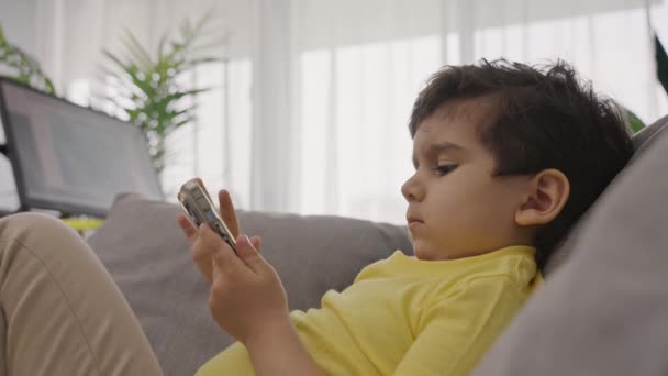 小さな甘い子供は 自宅でデジタルガジェットと ビデオゲームをプレイしながら 携帯電話のタッチスクリーン上の電子アプリケーションを使用してソファの上でリラックス — ストック動画