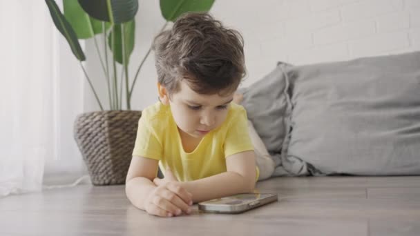 かわいい子供の男の子は 携帯電話の画面を見て 漫画を見て モバイルゲームをプレイし 屋内の床に横たわっています 携帯電話を使用して現代の技術の子供に適応 — ストック動画