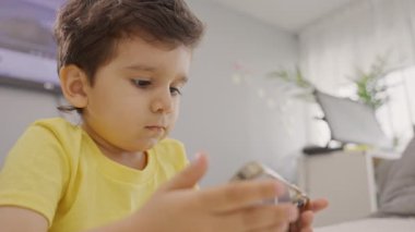 Anaokulu çocuğu sarı tişörtlü, elinde telefon, mobil uygulama kullanıyor, evde oyun oynuyor, cep telefonu ekranına bakıyor, evdeki koltukta oturuyor.