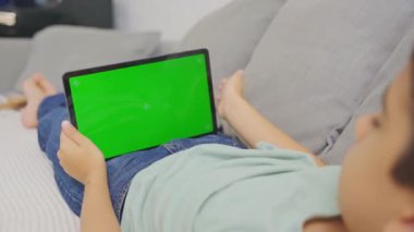 Çocuk kanepeye uzanmış, tableti tutuyor ve tıklıyor, bilgisayar yatay yeşil ekranlı..