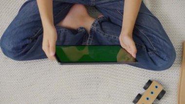 Çocuk kanepede oturuyor, sağa sola yatay yeşil ekranlı bir tablet eğiyor. Krom anahtar. Üst görünüm