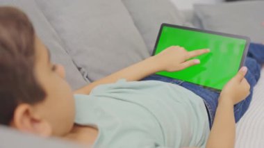 Çocuk kanepeye uzanmış, tableti tutuyor ve tıklıyor, bilgisayar yatay yeşil ekranlı..
