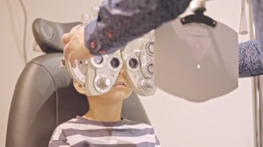 Küçük çocuklar göz muayenesi için göz doktoruna gittiklerinde phoropter kullanmışlar. Doktor elleri phoropter 'i genç çocuğun gözlerine yaklaştırıyor.